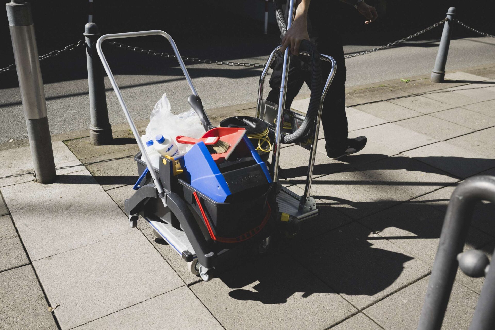 Reinigungswagen mit Putzutensilien und Eimer auf einem Gehweg bei Tageslicht.