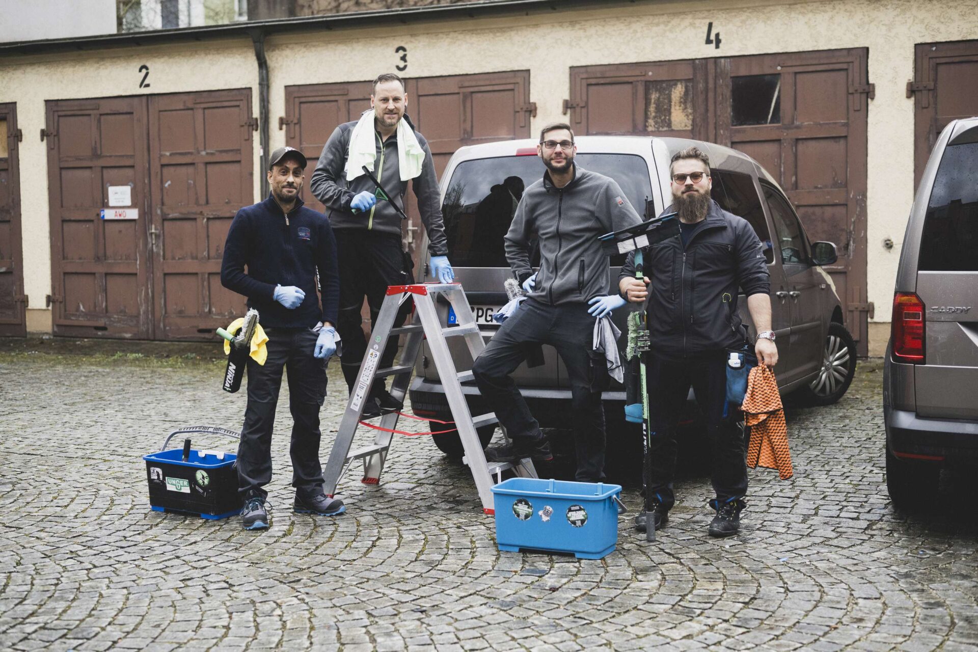 Vier Männer in Arbeitskleidung mit Reinigungsutensilien stehen vor einem Transporter auf einem gepflasterten Hof.
