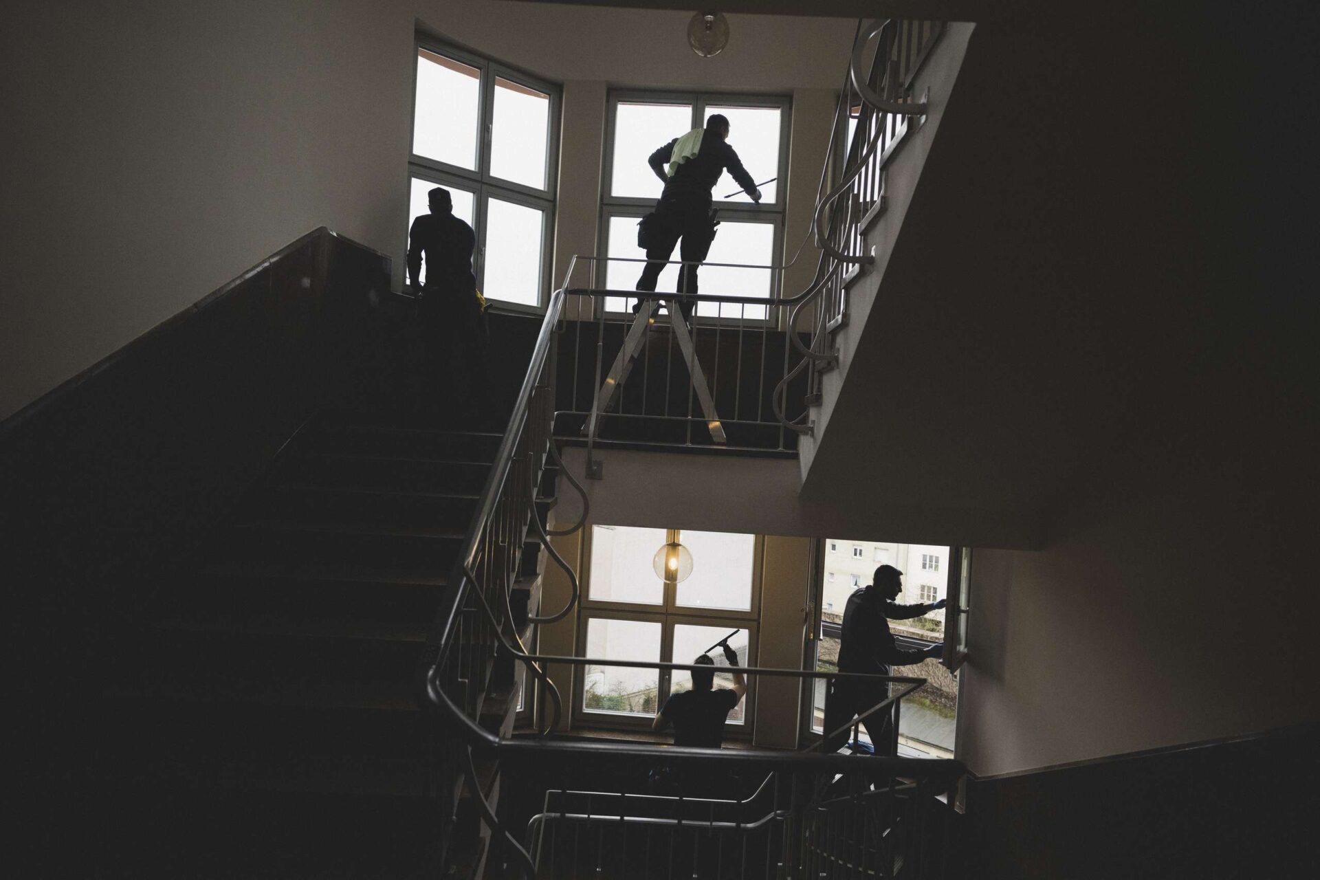 Vier Personen reinigen Fenster in einem Treppenhaus, Silhouetten vor großen Fenstern mit Tageslicht.