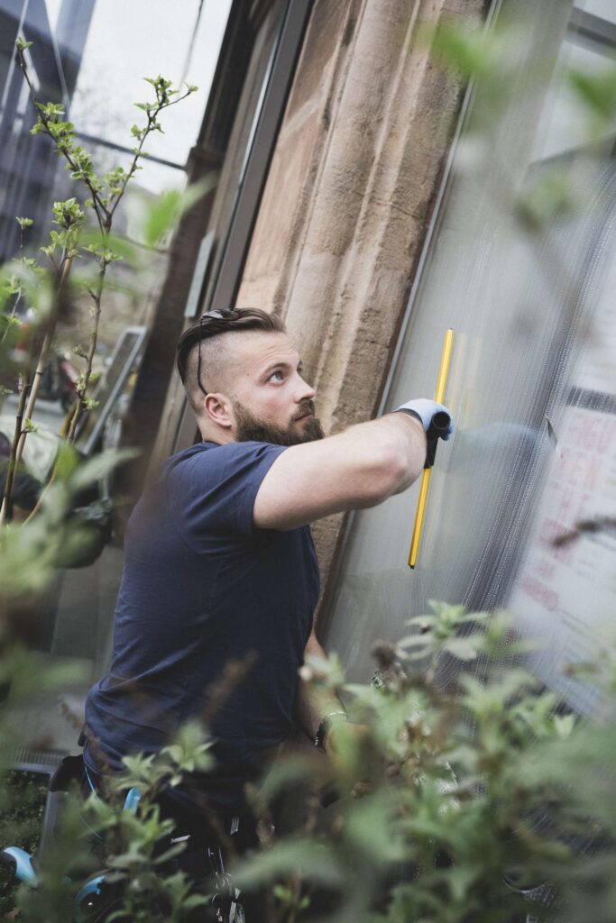 Mann mit Bart und Handschuhen reinigt mit einem Fensterwischer eine große Fensterscheibe von außen.