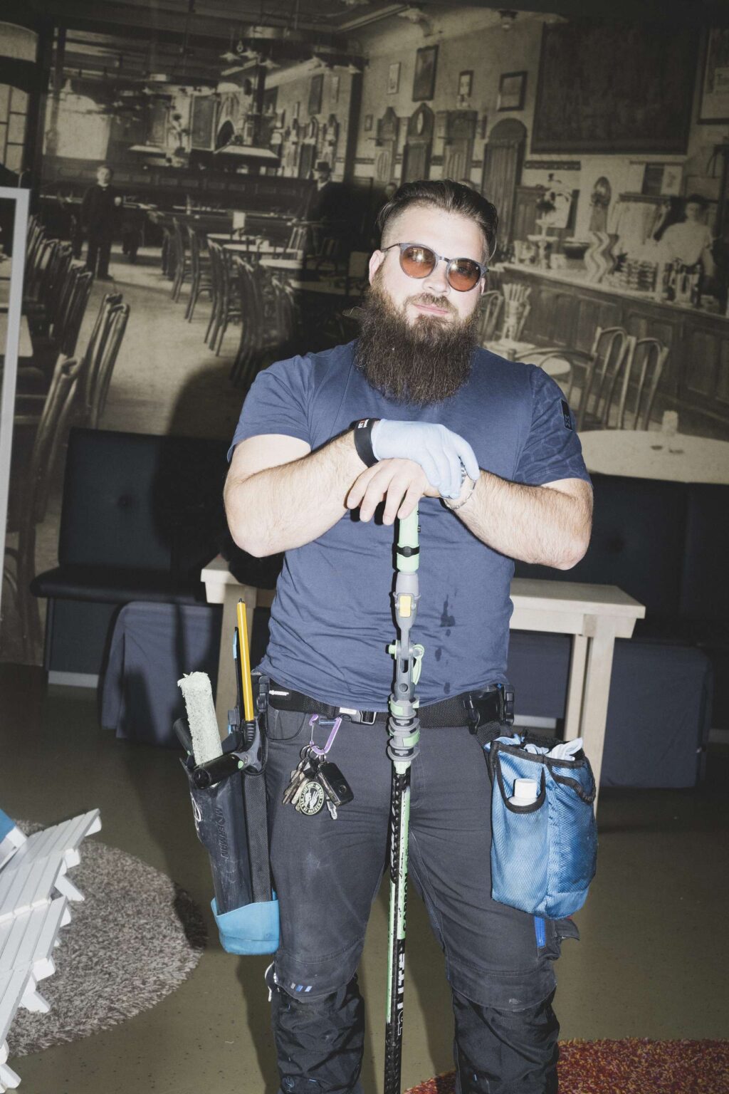 Mann mit Bart und Sonnenbrille steht mit Reinigungswerkzeug und Teleskopstange in einem Innenraum, trägt Handschuhe und Arbeitskleidung.