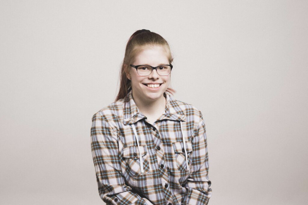 Junge Frau mit Brille und kariertem Hemd sitzt lächelnd vor hellem Hintergrund.