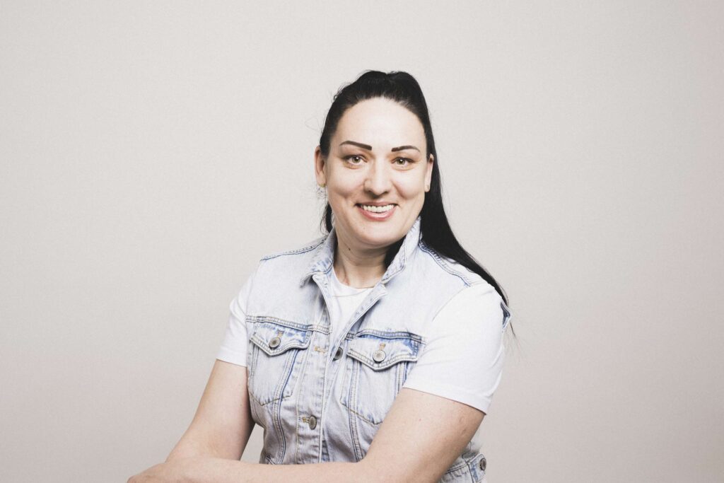 Frau mit langen dunklen Haaren, weißem T-Shirt und ärmelloser Jeansweste, lächelt in die Kamera vor hellem Hintergrund.