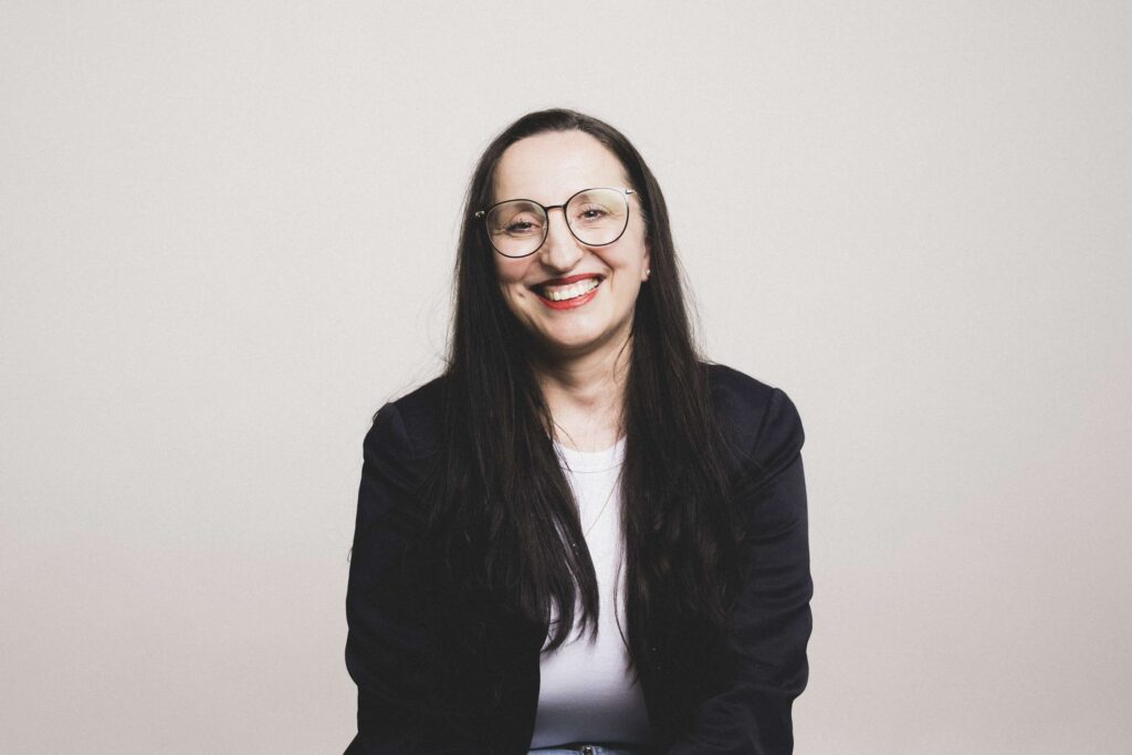 Frau mit langen dunklen Haaren, Brille und dunklem Blazer sitzt lächelnd vor hellem Hintergrund.