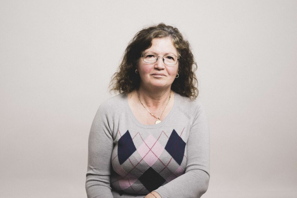 Frau mit lockigen, schulterlangen Haaren, Brille und hellem Pullover mit Rautenmuster sitzt vor hellem Hintergrund.