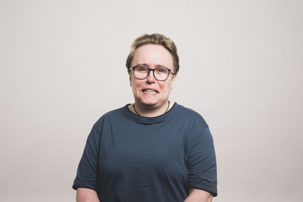 Frau mit kurzem, hellbraunem Haar und Brille, frontal vor hellem Hintergrund, trägt ein dunkelblaues T-Shirt und lächelt leicht.
