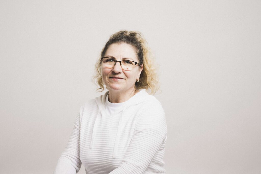 Frau mit lockigem, blondem Haar, Brille und weißem Kapuzenpullover vor hellem Hintergrund, Blick in die Kamera, dezentes Lächeln.