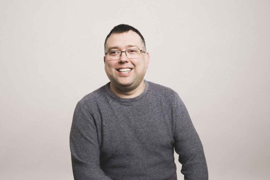 Mann mit Brille und grauem Pullover sitzt vor hellem Hintergrund und lächelt in die Kamera.