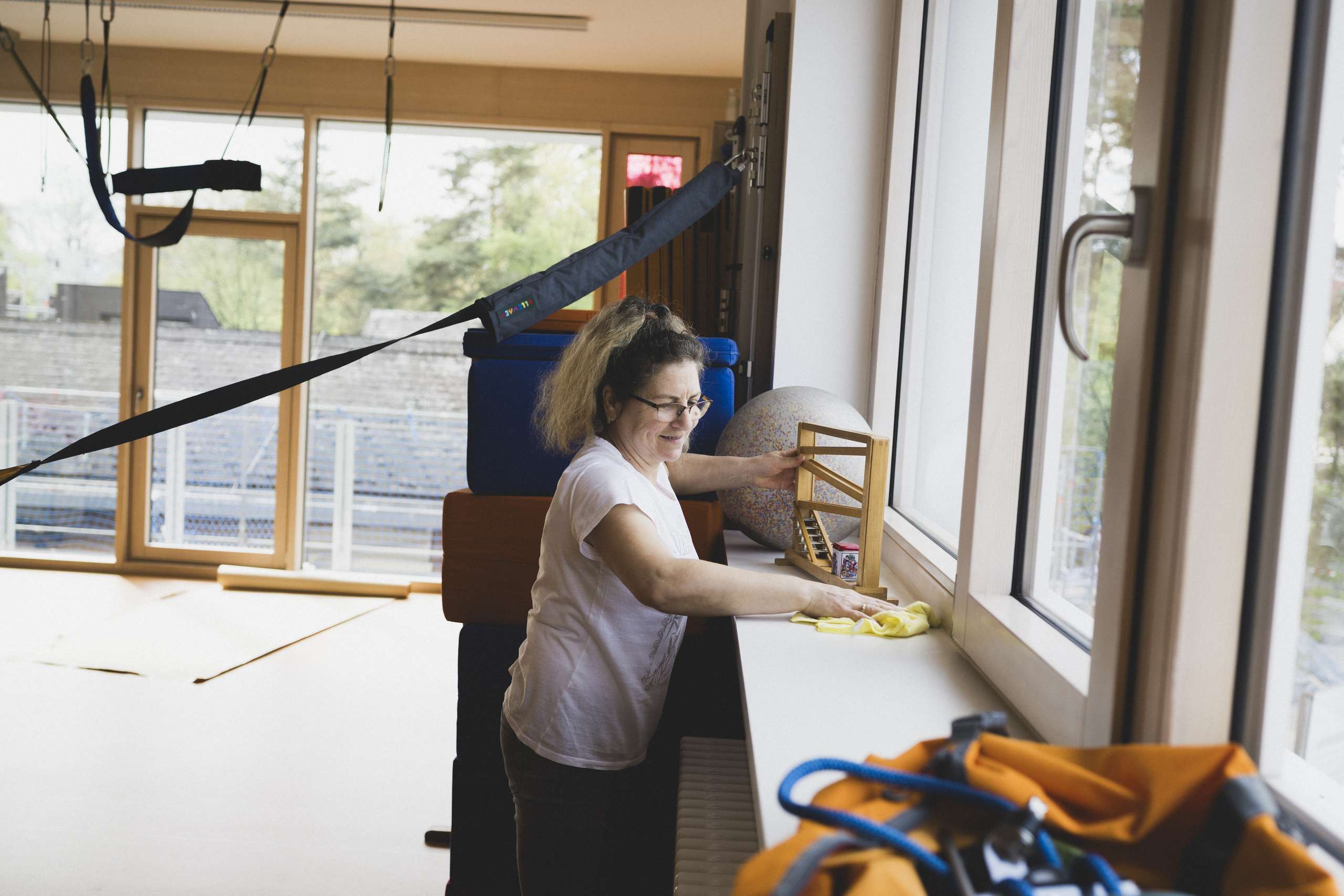 Frau mit Brille und hellem Oberteil wischt mit einem gelben Tuch eine Fensterbank in einem hellen Raum mit Sportgeräten ab.