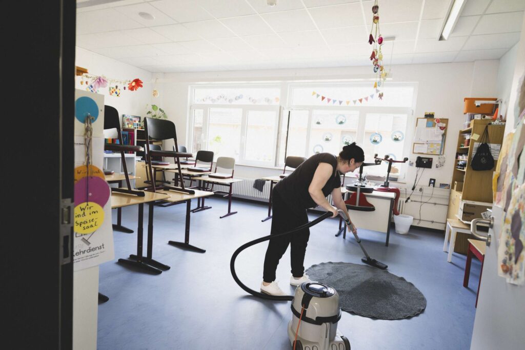 Frau mit Staubsauger reinigt einen Teppich in einem hellen Klassenzimmer mit hochgestellten Stühlen.