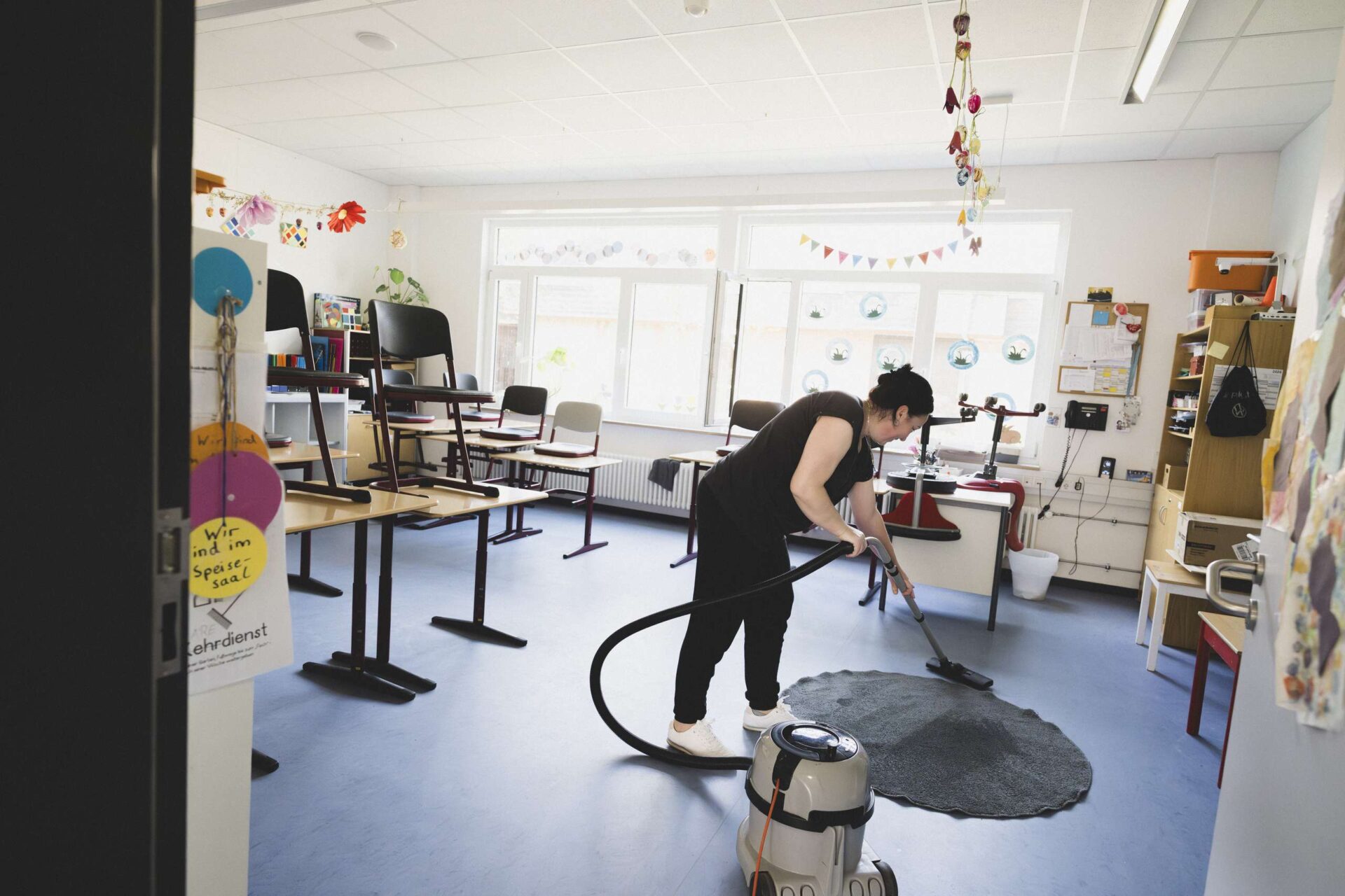 Frau mit Staubsauger reinigt einen Teppich in einem hellen Klassenzimmer mit hochgestellten Stühlen.
