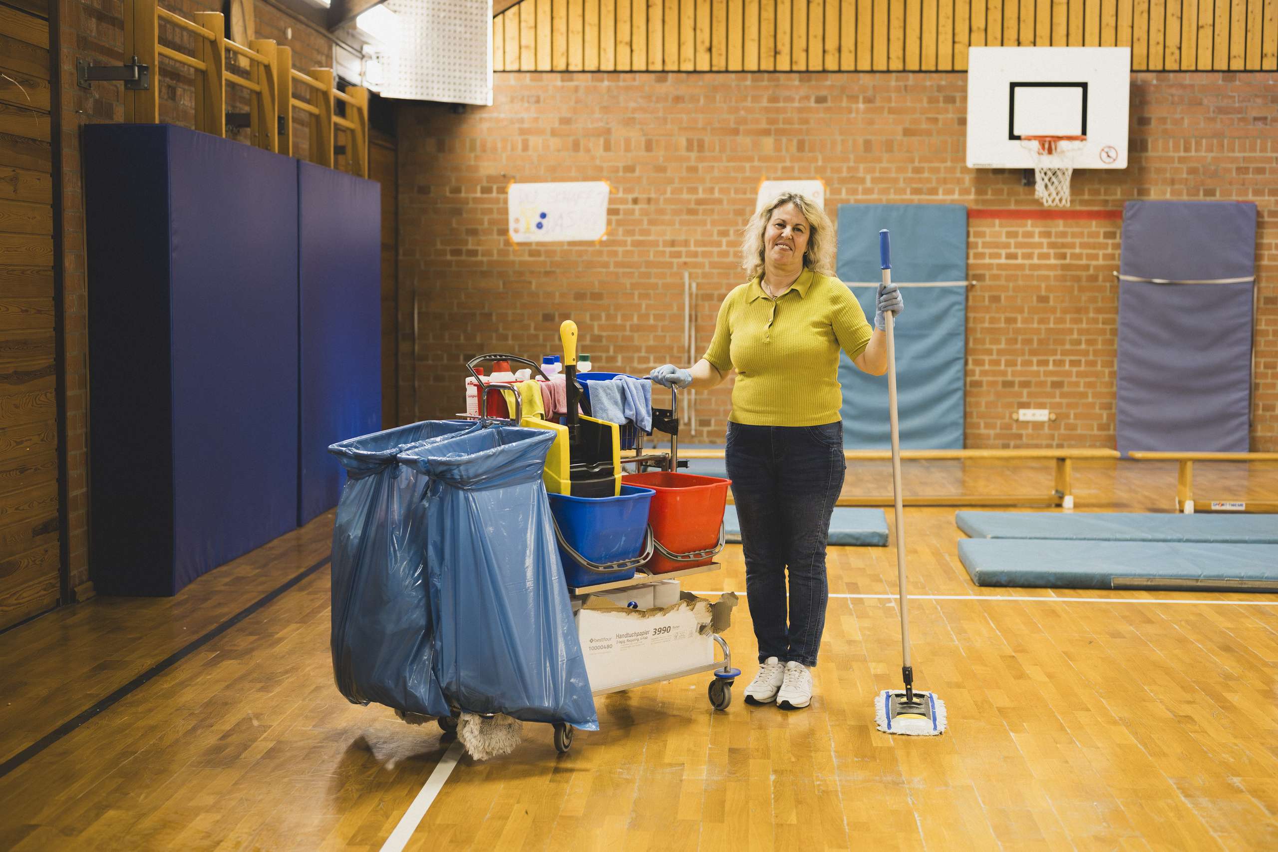Frau mit blondem Haar steht in Sporthalle neben Reinigungswagen mit mehreren Eimern und Müllsäcken, hält einen Wischmopp in der Hand.