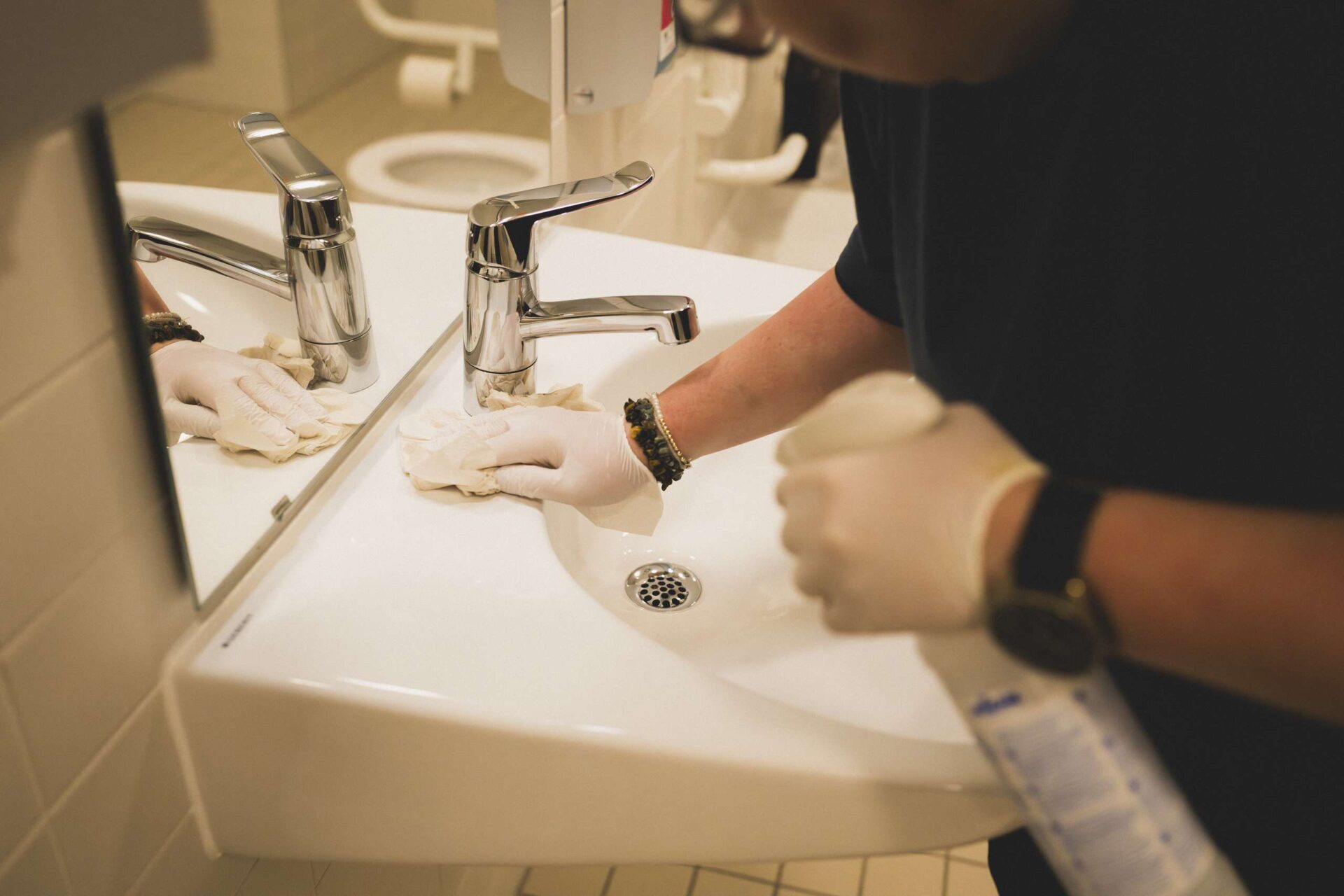 Person mit Einweghandschuhen reinigt ein weißes Waschbecken mit einem Tuch in einem Badezimmer.