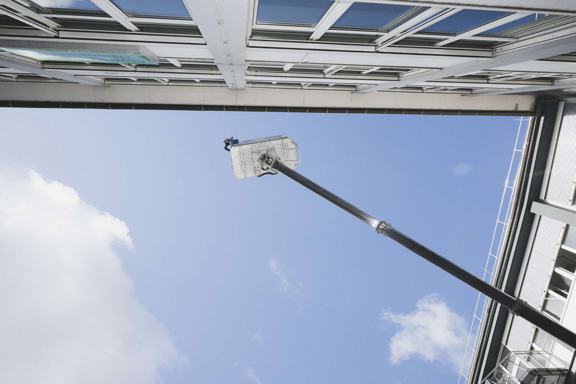 Arbeitsbühne mit Person in großer Höhe an Glasfassade eines modernen Gebäudes vor blauem Himmel.