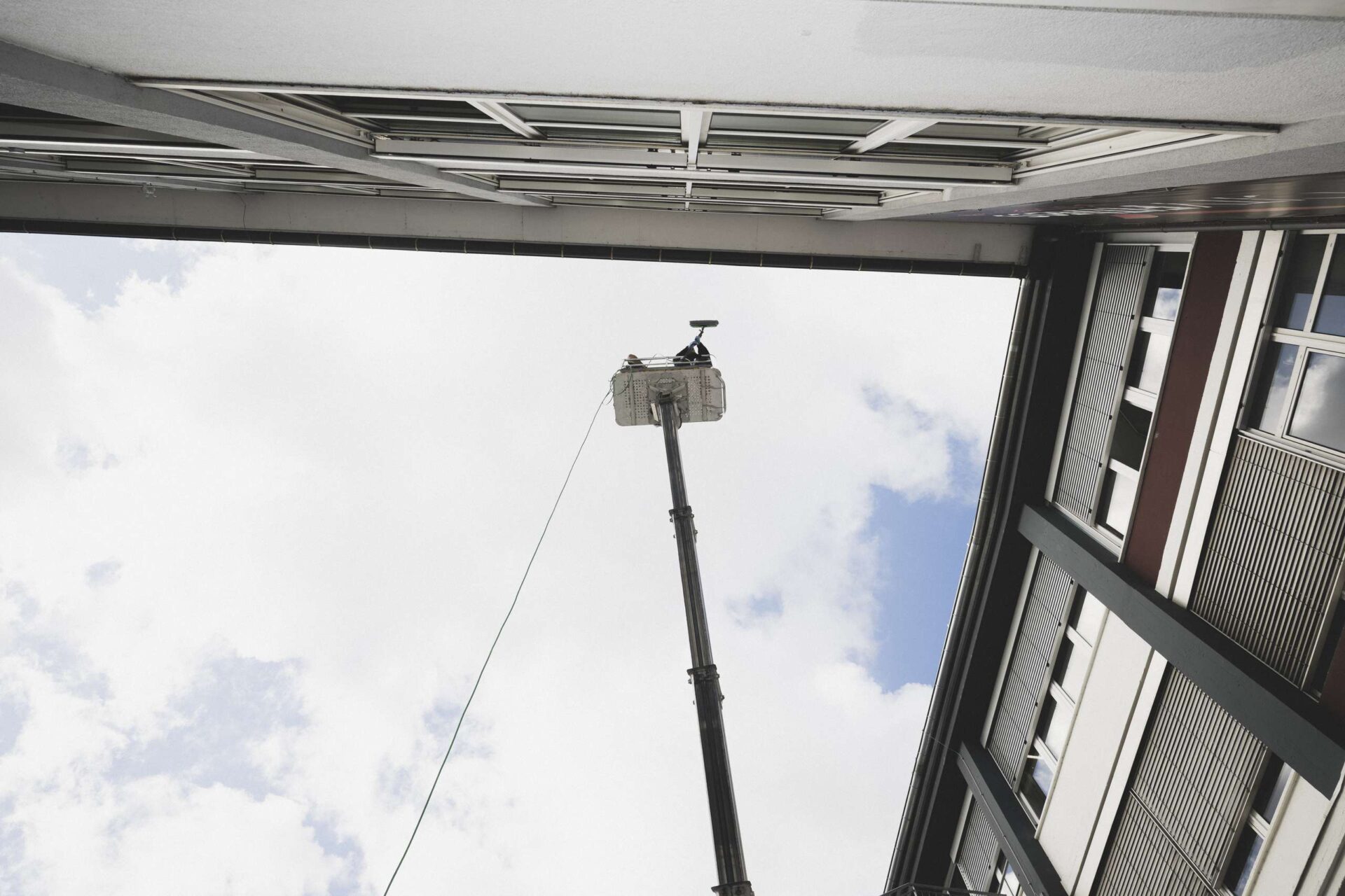 Person in Arbeitskleidung steht auf einer Hebebühne und reinigt von außen die Fenster eines mehrstöckigen Gebäudes.
